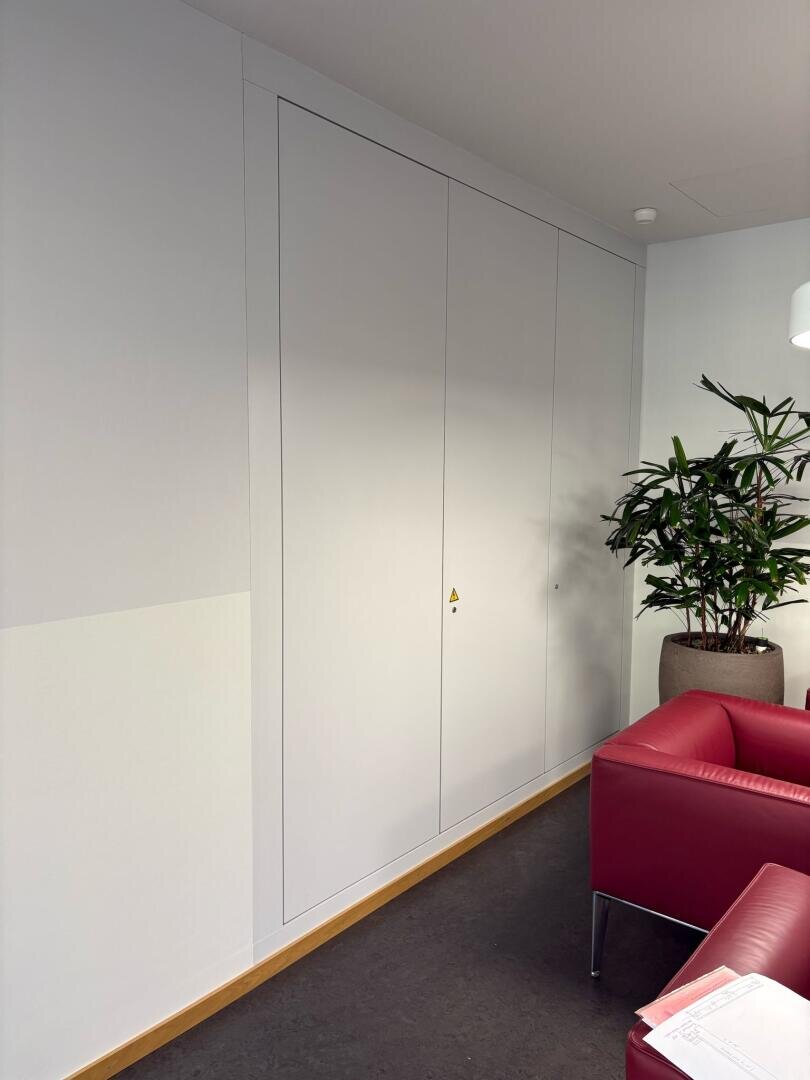Ein großer, weißer Einbauschrank mit geschlossenen Türen steht vor einer hellen Wand. In der Nähe stehen eine Topfpflanze und ein roter Sessel, auf dessen Armlehne ein Stück Papier liegt.