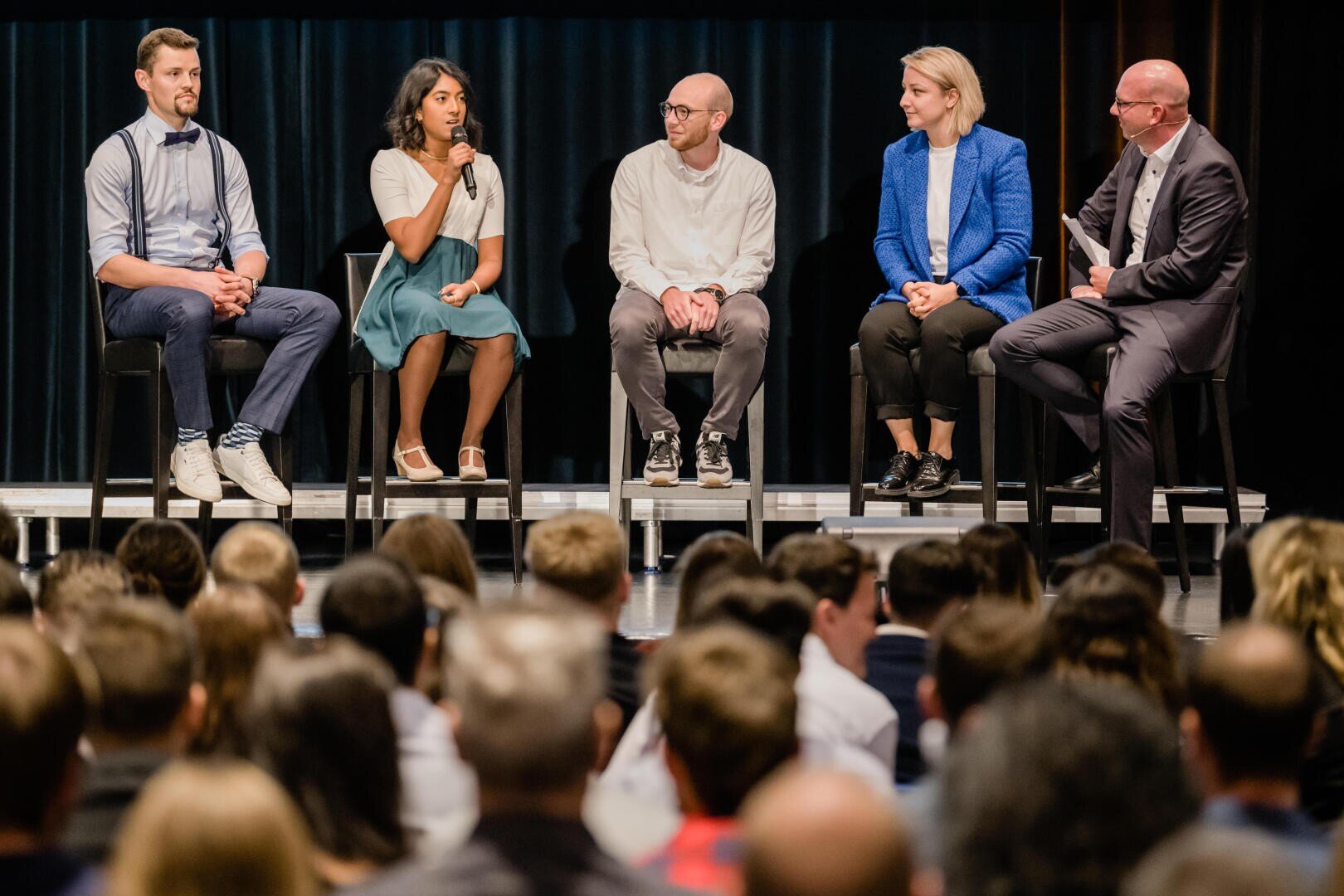Fünf Personen sitzen während einer Podiumsdiskussion vor einem Publikum auf einer Bühne. Eine Person hält ein Mikrofon und spricht, während die anderen zuhören. Alle sind in legerer Business-Kleidung gekleidet.
