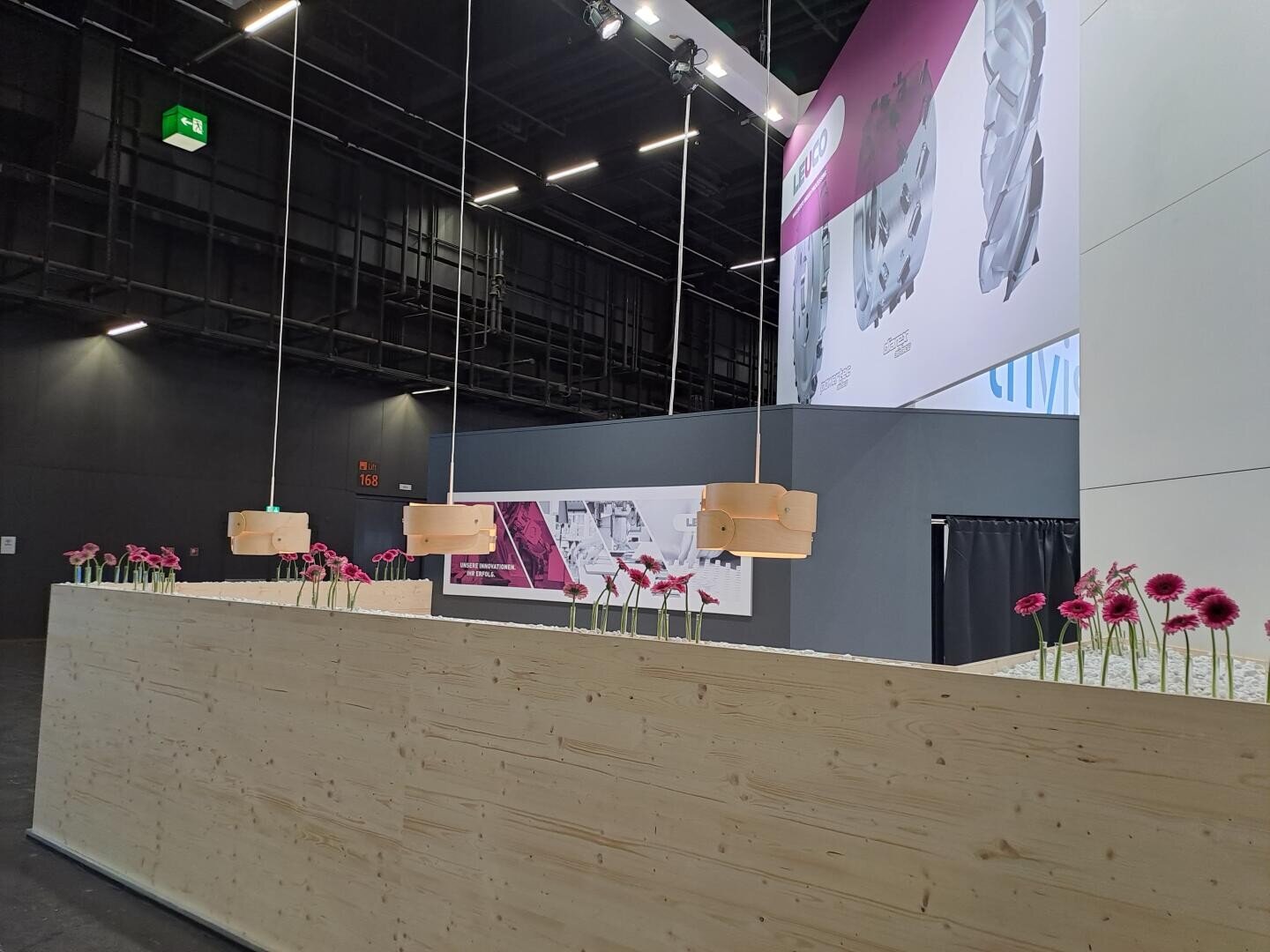 Un intérieur moderne avec un long comptoir en bois décoré de fleurs roses réparties uniformément dans des vases, des lampes suspendues au-dessus et des plafonds sombres de style industriel. En arrière-plan, de grands posters et une cloison grise.