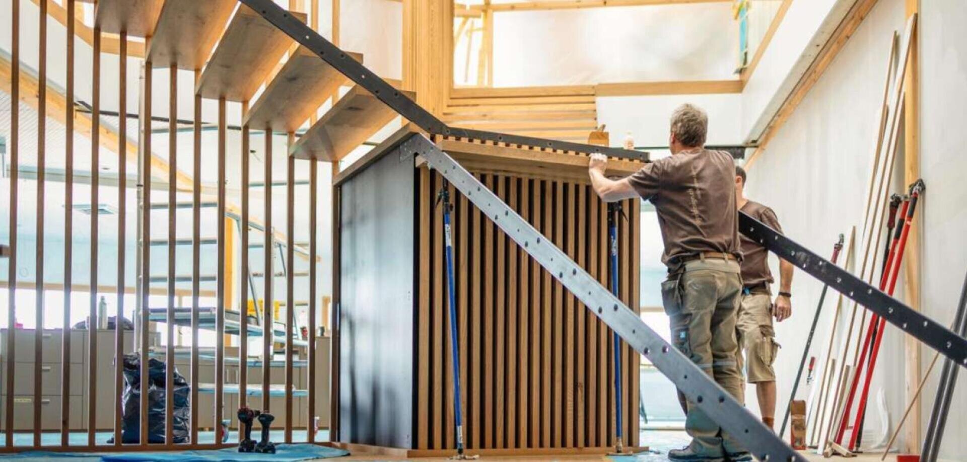 Zwei Personen bauen in einem Innenraum eine moderne Holzstruktur zusammen, umgeben von Bauwerkzeugen und -materialien, mit einer teilweise fertigen Treppe und vertikalen Holzlatten im Hintergrund.