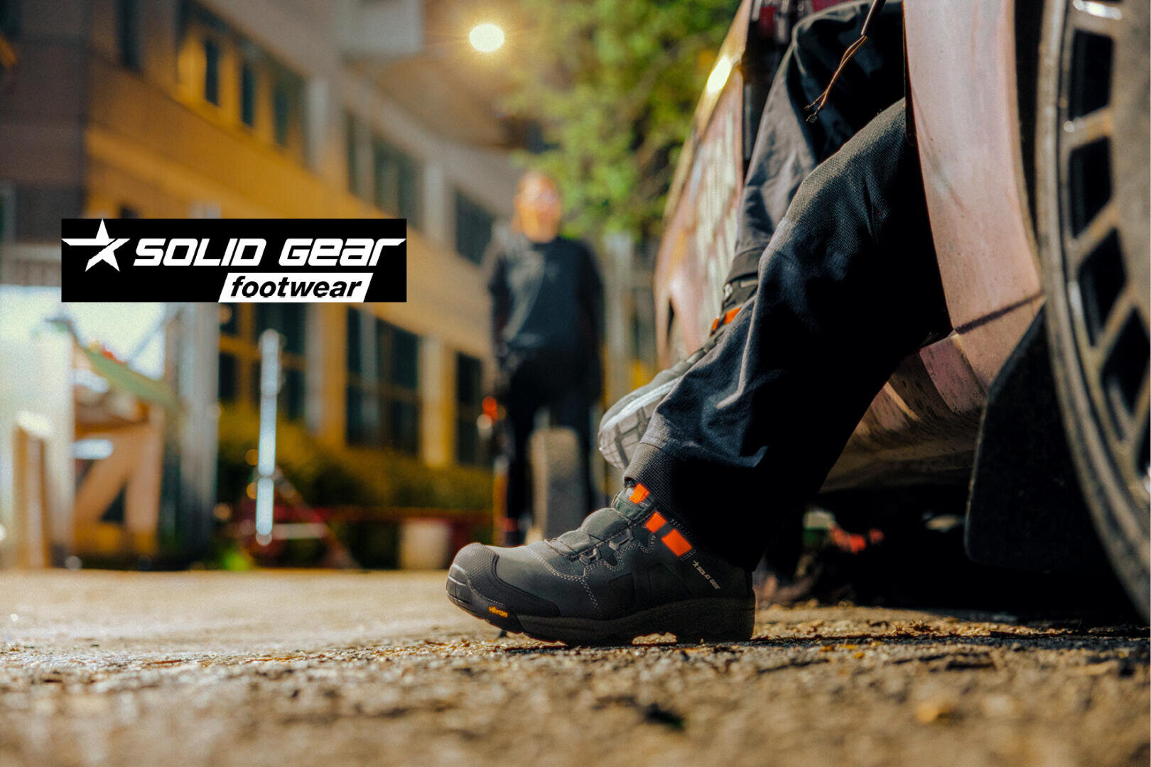 Une personne portant des chaussures noires Solid Gear est assise de nuit sur le bord d'un véhicule, tandis qu'un autre personnage flou se tient sur une route en arrière-plan. Le logo de Solid Gear Footwear apparaît à gauche.