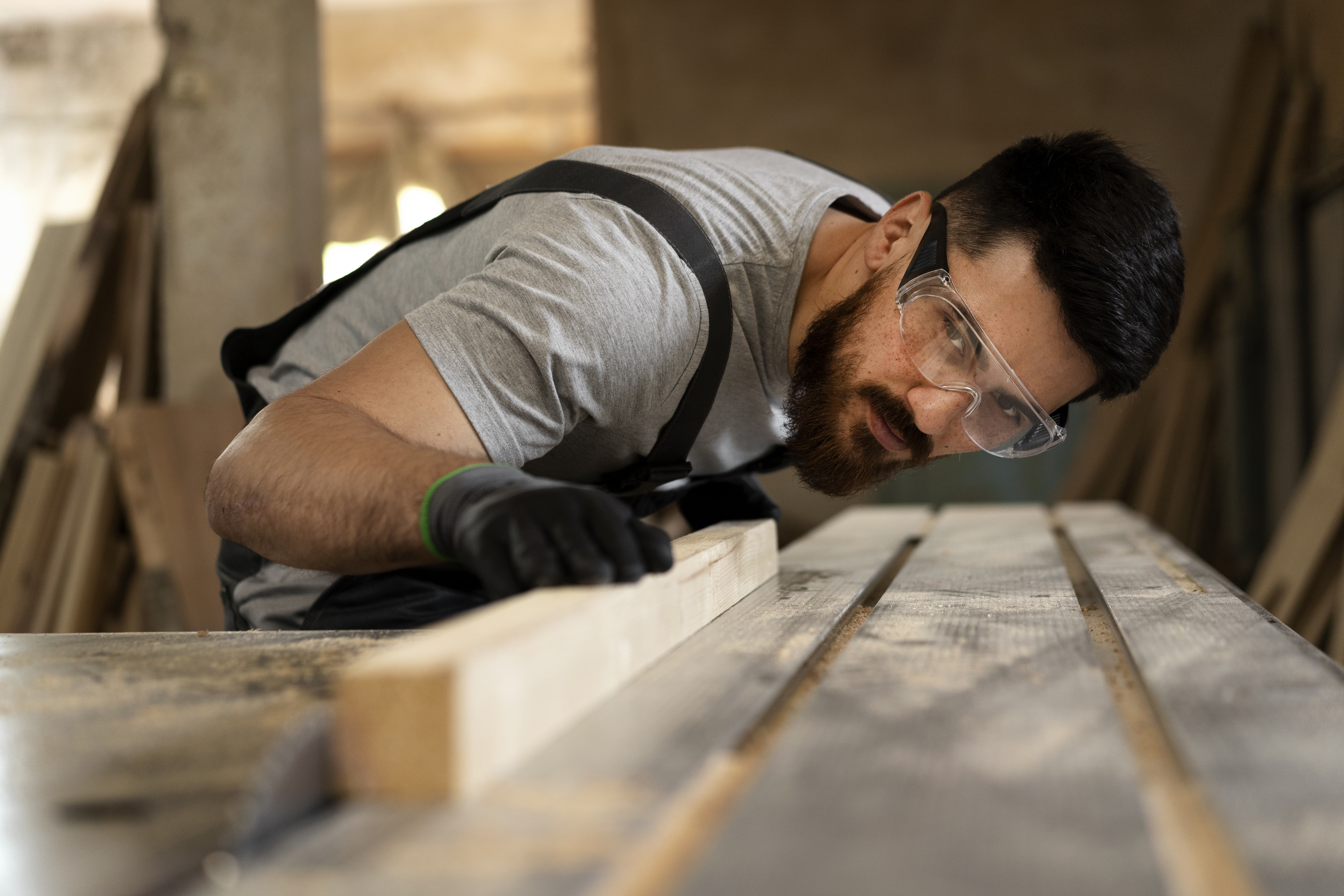 Ein bärtiger Mann mit Schutzbrille und Handschuhen prüft in einer Werkstatt ein Holzbrett, während er sich über eine Werkbank beugt, die von Holzbearbeitungswerkzeugen und Sägemehl umgeben ist.