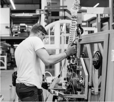 Eine Person, die eine Schutzbrille trägt, bedient eine große Industriemaschine in einer Fabrik, umgeben von Maschinen und teilweise montierten Fensterrahmen.