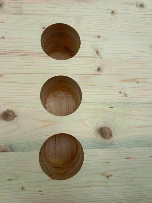 Eine helle Holzoberfläche mit drei gleichmäßig verteilten, runden Löchern, die in einer vertikalen Linie gebohrt sind. Das Holz weist sichtbare Maserungen und Äste auf.