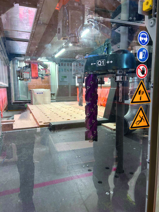 Eine CNC-Maschine mit einem violetten Bohrer ist hinter einer Sicherheitsglasscheibe mit Warn- und Sicherheitszeichen in Betrieb. Im Inneren des Arbeitsraums sind Holzteile und industrielle Geräte zu sehen.
