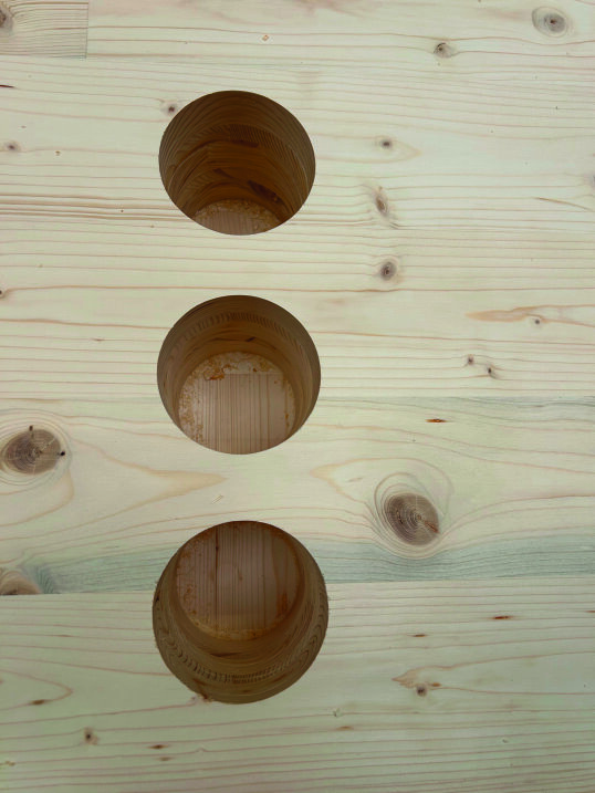 Une planche en bois clair avec trois grands trous circulaires régulièrement espacés, percés verticalement en rangée à travers la surface. Les veines et les nœuds du bois sont visibles sur toute la planche.