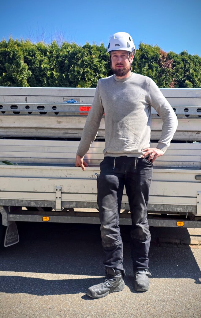 Ein Mann, der einen weißen Schutzhelm, ein hellgraues langärmeliges Hemd, eine schwarze Hose und Arbeitsstiefel trägt, steht vor einem mit Metallleitern beladenen Lastwagen. Im Hintergrund sind grüne Büsche und blauer Himmel zu sehen.