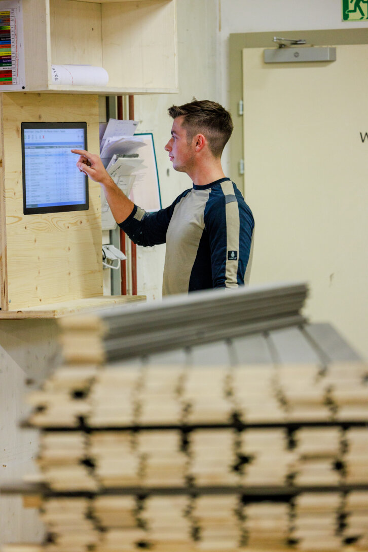 Ein Mann steht vor einem an der Wand montierten Touchscreen und interagiert mit einer digitalen Anzeige, während er Papiere in der Hand hält; gestapelte Holzbretter sind im Vordergrund verschwommen.