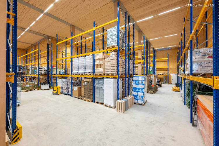 Un entrepôt spacieux avec de hautes étagères en métal bleu et jaune stockant des palettes de boîtes, de colis et de matériaux de construction, le tout soigneusement disposé en rangées sous un plafond en bois brillamment éclairé.