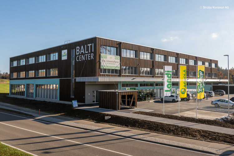 Un bâtiment moderne de trois étages, orné de l'inscription « BALTI CENTER » et de drapeaux Landi verts et jaunes. Il est doté de grandes fenêtres et de boiseries. Un parking et une rue sont visibles au premier plan.
