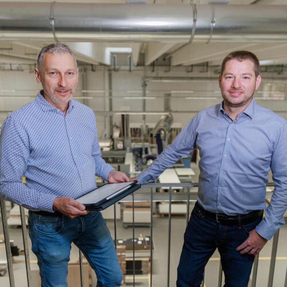 Zwei Männer in legeren Button-down-Hemden und Jeans stehen drinnen an einem Geländer, einer hält ein Tablet in der Hand. Im Hintergrund sind Industrieanlagen und Arbeitsbereiche zu sehen, die an eine moderne Fabrik oder Werkstatt erinnern.