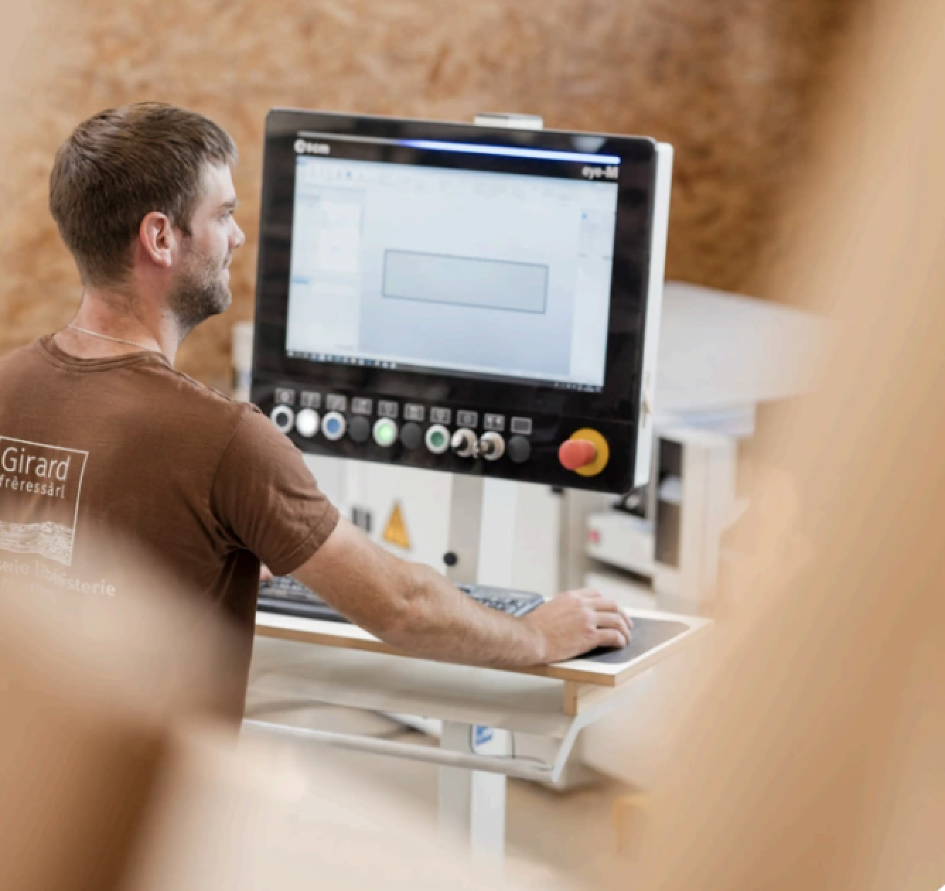 Eine Person sitzt an einer Kontrollstation und bedient einen großen Touchscreen, auf dem technische Diagramme angezeigt werden. Die Person trägt ein braunes Hemd, im Hintergrund sind Holzoberflächen und unscharfe Objekte zu sehen.