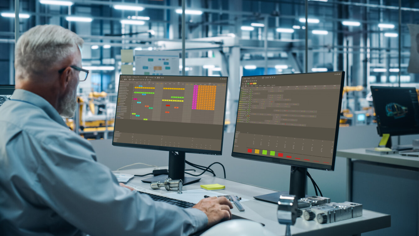 Eine Person mit grauen Haaren und Bart arbeitet an einem Schreibtisch mit zwei großen Monitoren, auf denen bunte Diagramme und Daten in einer modernen, hochtechnisierten Büro- oder Fabrikumgebung angezeigt werden.