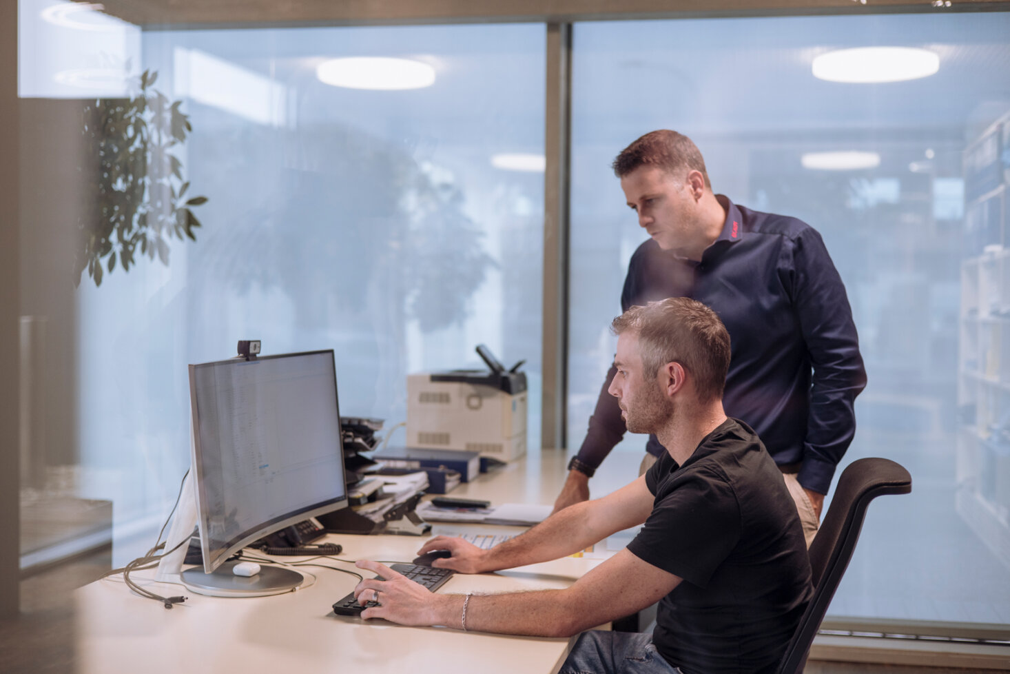 Zwei Männer arbeiten in einem Büro gemeinsam an einem Computertisch; einer sitzt und bedient die Tastatur, während der andere daneben steht und auf den Monitor schaut. Das Büro hat große Fenster und ist modern eingerichtet.