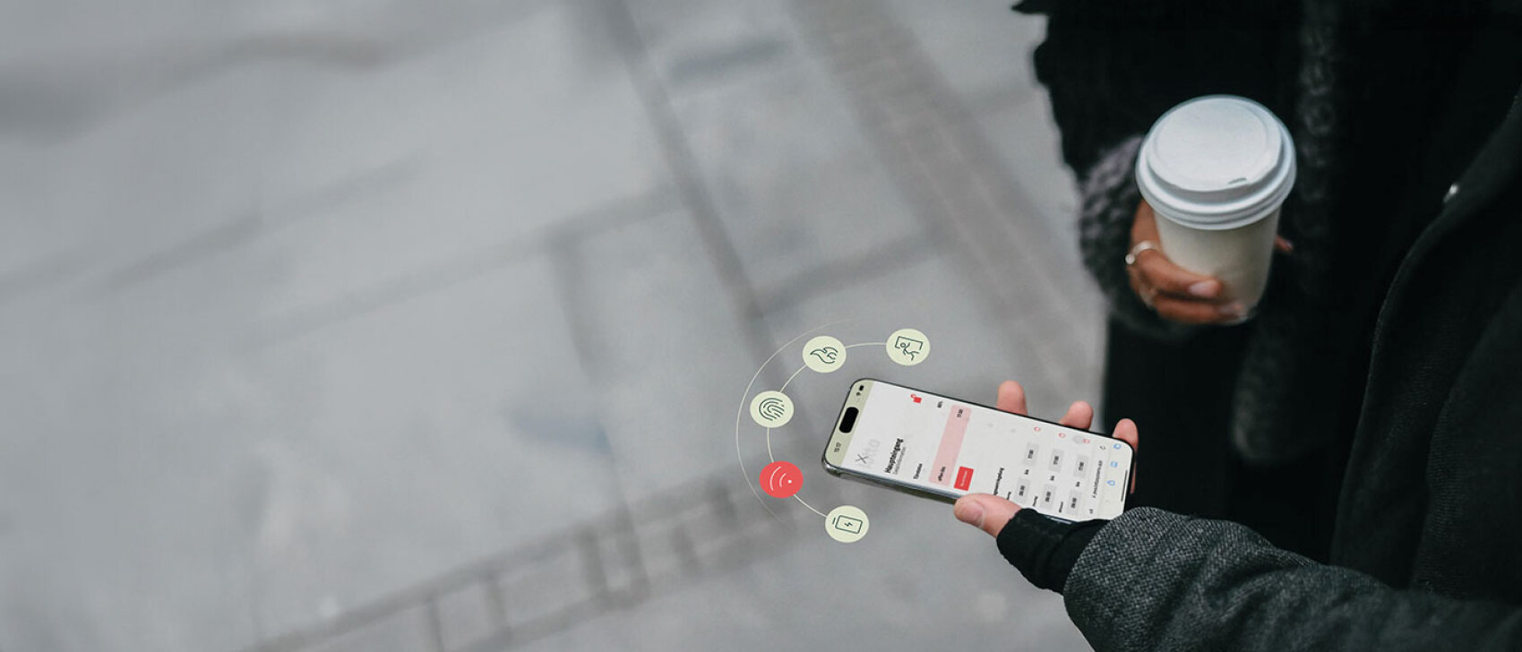 Eine Person in einem dunklen Mantel hält eine Kaffeetasse in der einen und ein Smartphone in der anderen Hand. Über dem Telefondisplay erscheinen digitale Symbole, die die Konnektivität oder die Nutzung einer App anzeigen. Der Hintergrund ist ein grauer, verschwommener Bürgersteig.