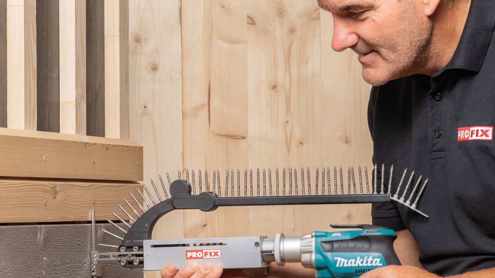 Ein Mann verwendet ein Makita-Elektrowerkzeug mit einem PROFIX-Aufsatz, um eine Reihe von Nägeln oder Schrauben in eine Holzterrasse einzubringen. Im Hintergrund sind helle Holzplatten zu sehen.