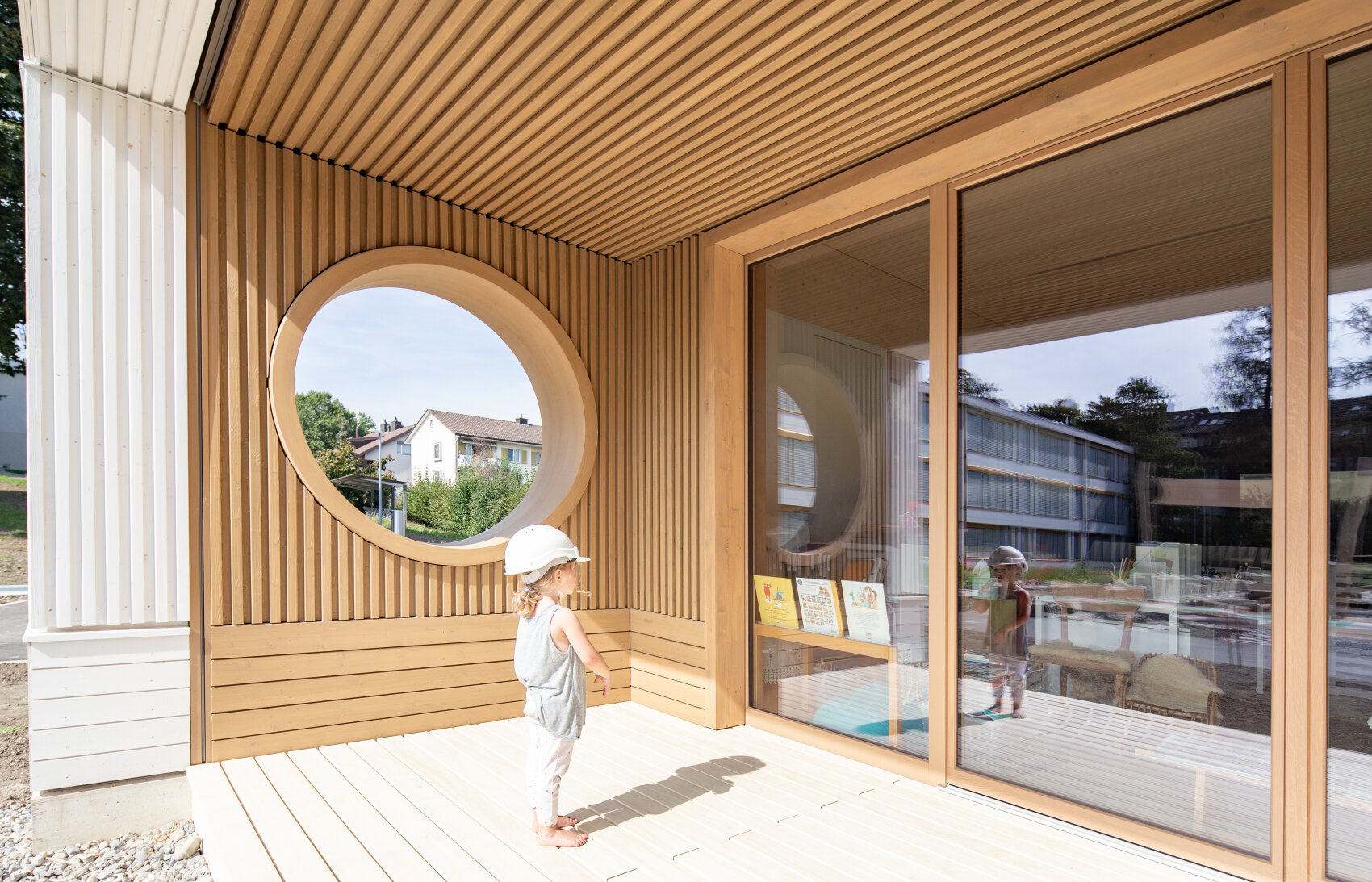 Un jeune enfant portant une tenue blanche et un casque se tient sous un porche en bois à lattes verticales, à côté d'une grande fenêtre ronde et de portes vitrées reflétant l'enfant. Des bâtiments modernes et de la verdure sont visibles à l'extérieur.