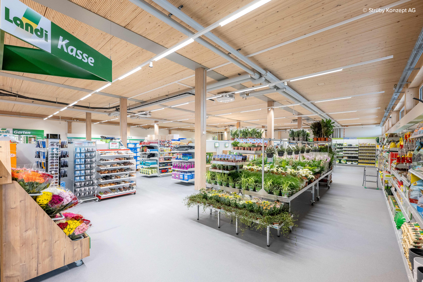 Un intérieur de magasin spacieux et bien éclairé avec des étagères remplies de produits variés, un étalage de plantes en pot et de fleurs au centre et un panneau vert indiquant « Caisse enregistreuse » suspendu au plafond.