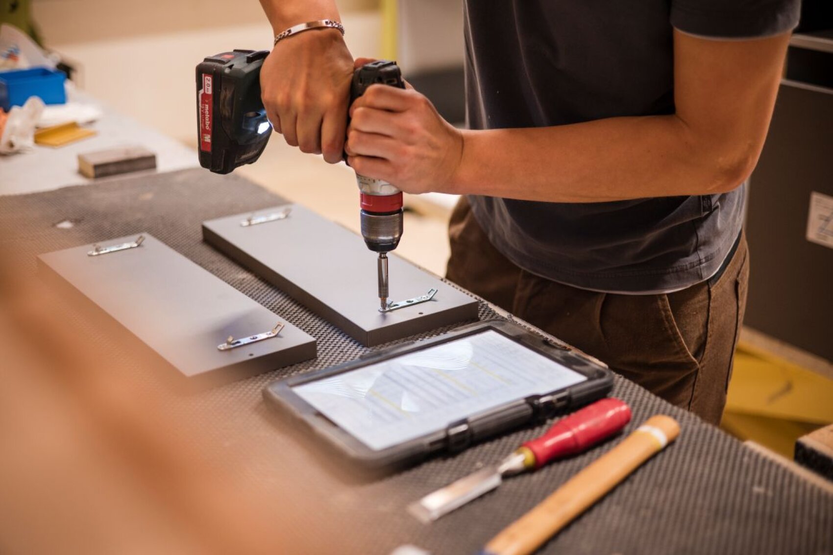 Une personne utilise une perceuse électrique pour fixer des charnières métalliques à un panneau de bois gris sur un établi, avec une tablette, un tournevis et d'autres outils à proximité.