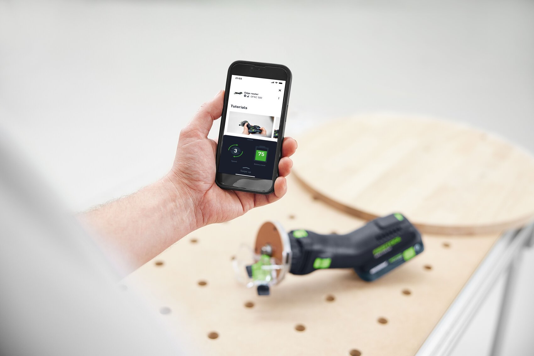 Une personne tient un smartphone avec une application d'outillage dans la main, tandis qu'un outil électrique noir et vert est posé sur une planche de bois ronde avec des trous dans un espace de travail lumineux.
