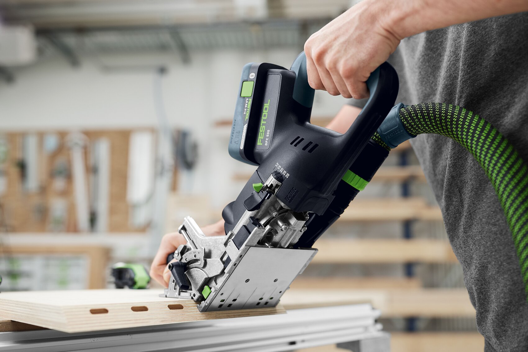 Eine Person verwendet ein Elektrowerkzeug mit einem grünen Schlauchaufsatz, um ein Stück helles Holz auf einer Werkbank in einer Werkstatt zu schneiden oder zu verbinden.