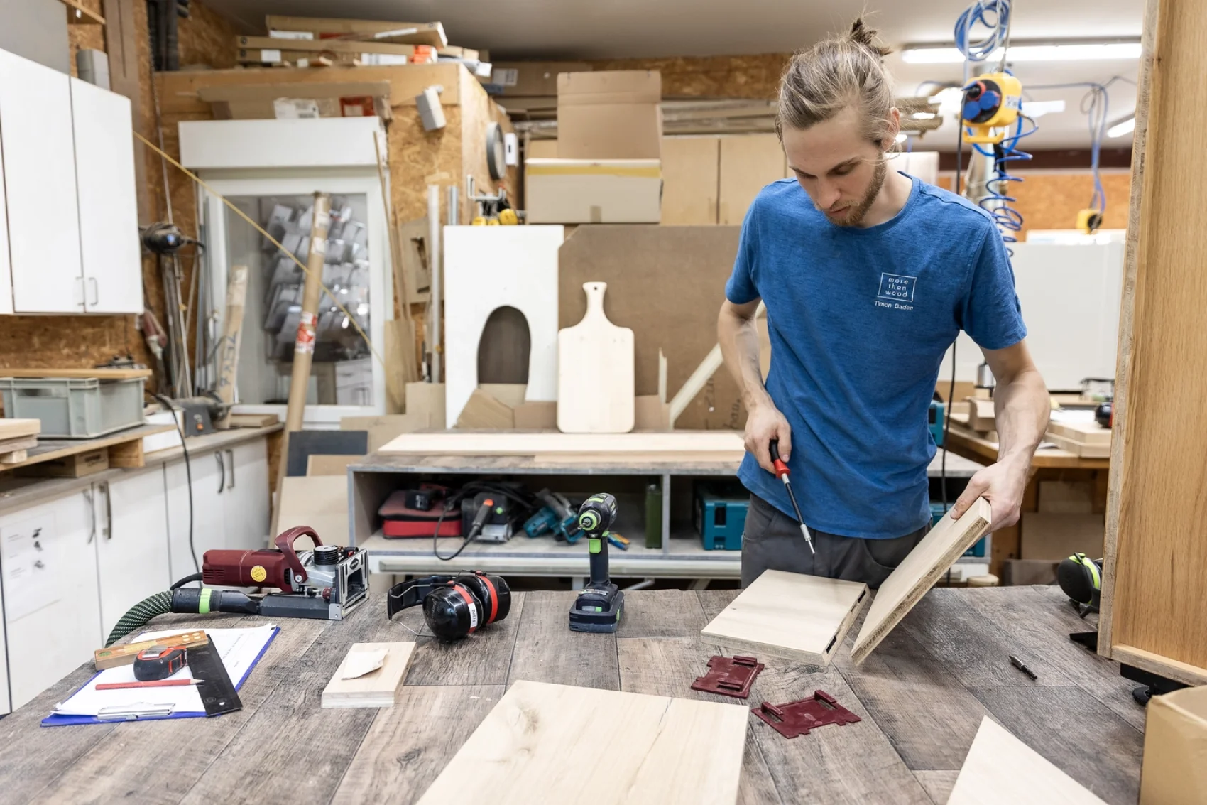 Ein Mann in einem blauen Hemd arbeitet in einer Werkstatt daran, Holzteile auf einer Werkbank zusammenzubauen. Werkzeuge, Holzplatten und Materialien sind um ihn herum verstreut und ein Briefbeschwerer liegt auf dem Tisch. Im Hintergrund befinden sich Regale und Schränke.
