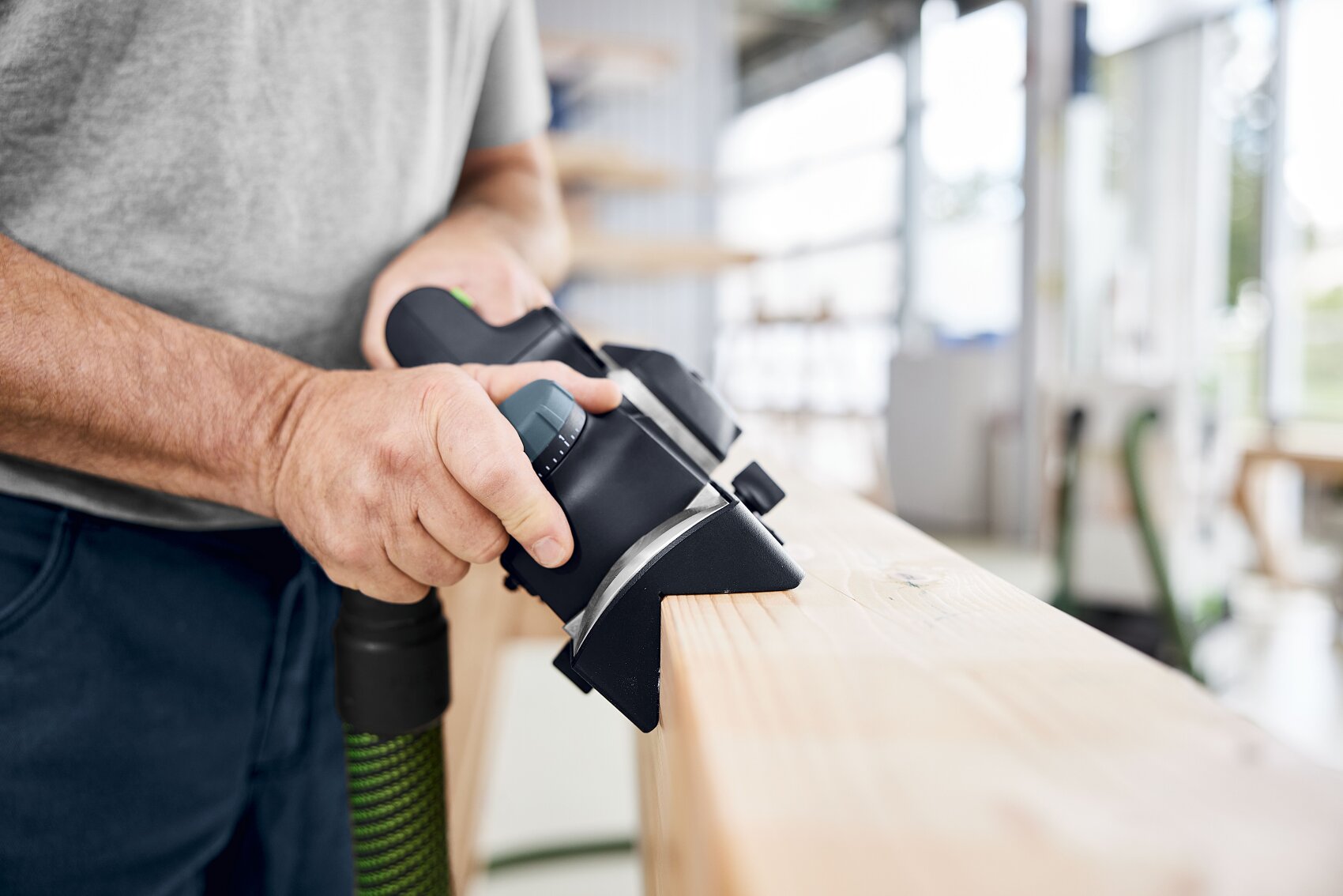 Dans un atelier bien éclairé, une personne lisse la surface d'une planche de bois avec une raboteuse électrique. Seuls ses mains et l'outil sont visibles.