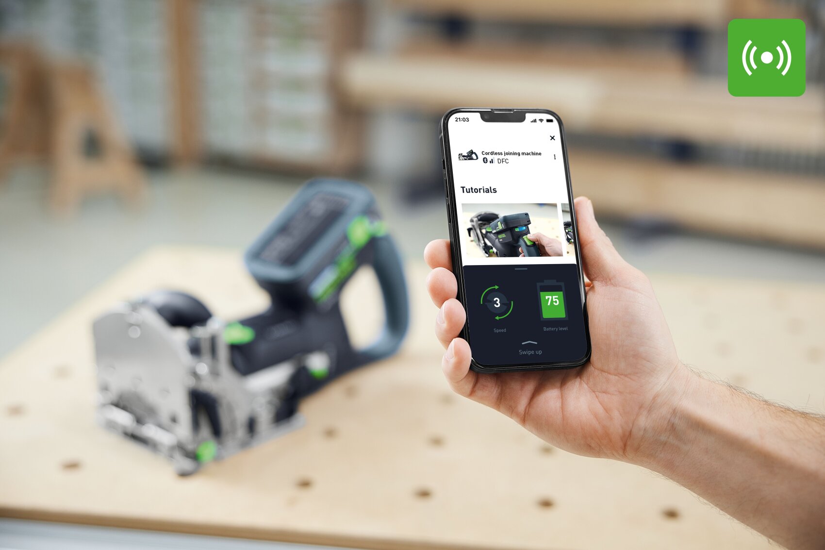 Eine Person hält ein Smartphone in der Hand, auf dem eine Anleitung für ein Elektrowerkzeug angezeigt wird, wobei das eigentliche Elektrowerkzeug auf einer Werkbank im Hintergrund verschwommen ist. Das grüne Funksignal-Symbol erscheint in der oberen rechten Ecke.