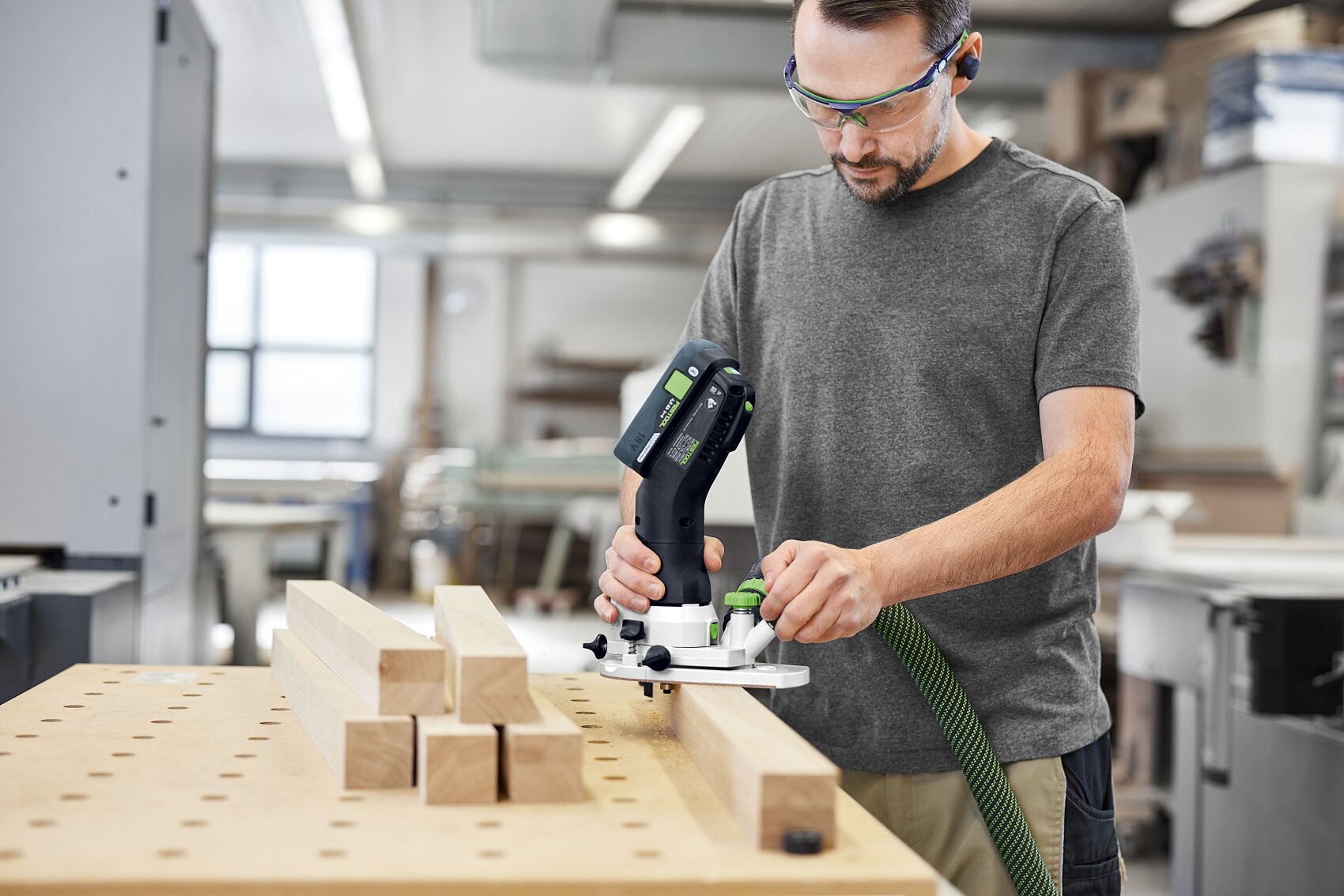 Un homme portant des lunettes de protection travaille avec un outil électrique sur une planche de bois dans un atelier. Plusieurs planches de bois sont empilées sur un établi à côté de lui. L'espace de travail est clair et bien rangé.