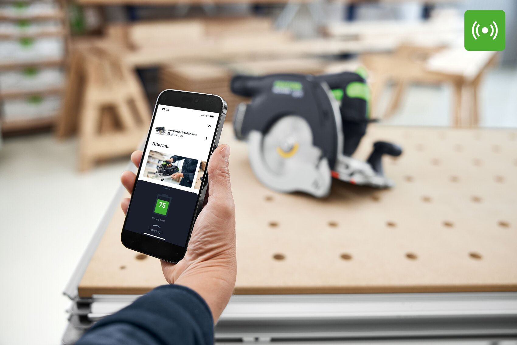 Une personne avec un smartphone affichant une application de travail du bois, avec une scie circulaire sur un établi perforé en arrière-plan flou ; une icône verte de signal radio apparaît dans le coin supérieur droit.