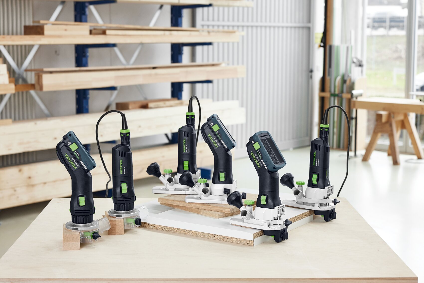 Cinq défonceuses électriques au design noir, vert et argenté sont disposées sur un établi dans un atelier de menuiserie lumineux avec des étagères, des planches de bois et de grandes fenêtres en arrière-plan.