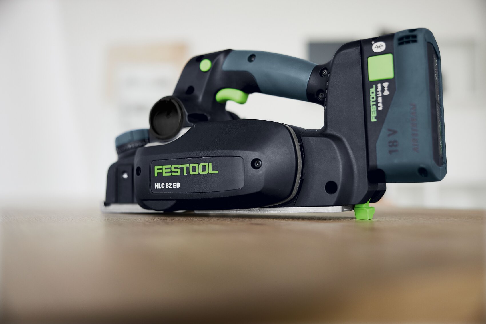 Un gros plan sur un rabot à main sans fil Festool HLC 82 EB, posé sur un support en bois. On peut voir le design noir et vert avec la marque visible et une batterie 18 V connectée.