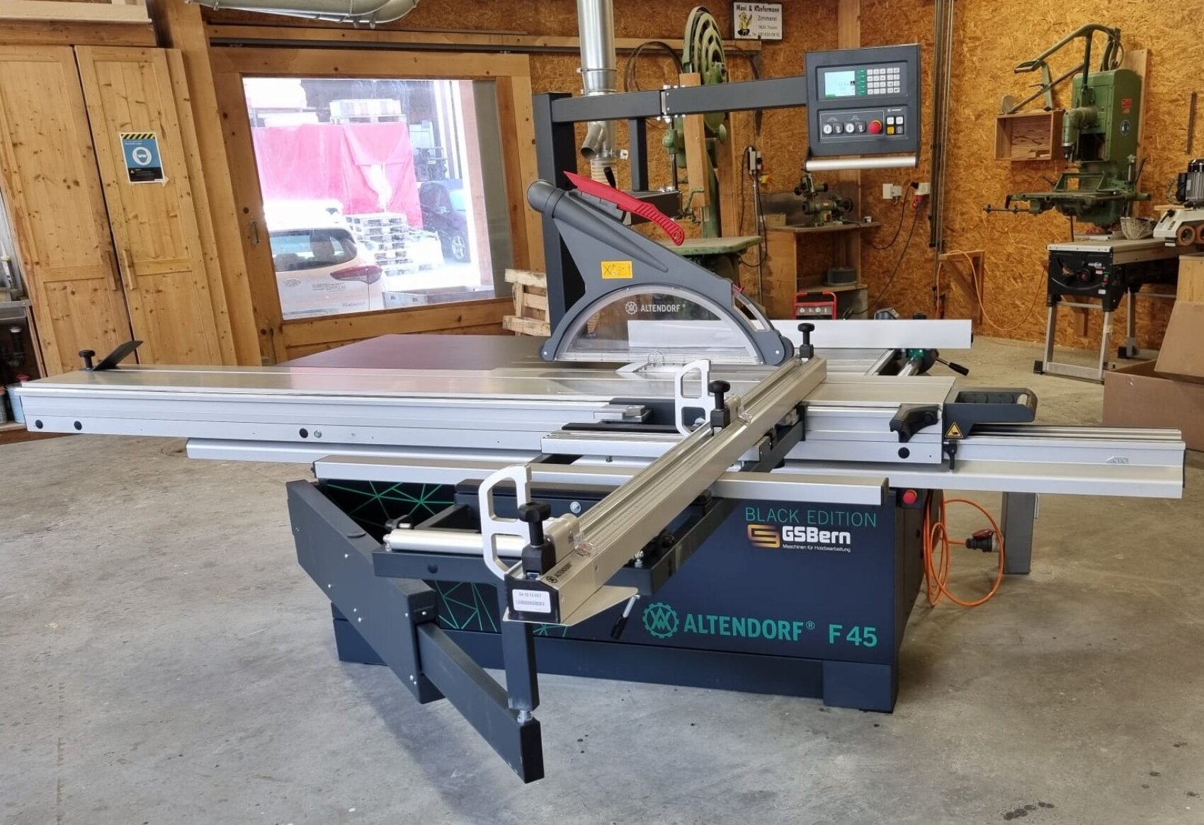 Eine große Altendorf F45 Formatkreissäge mit digitalem Bedienfeld steht in einer Holzwerkstatt, umgeben von Werkzeugen, Holzwänden und Schränken.