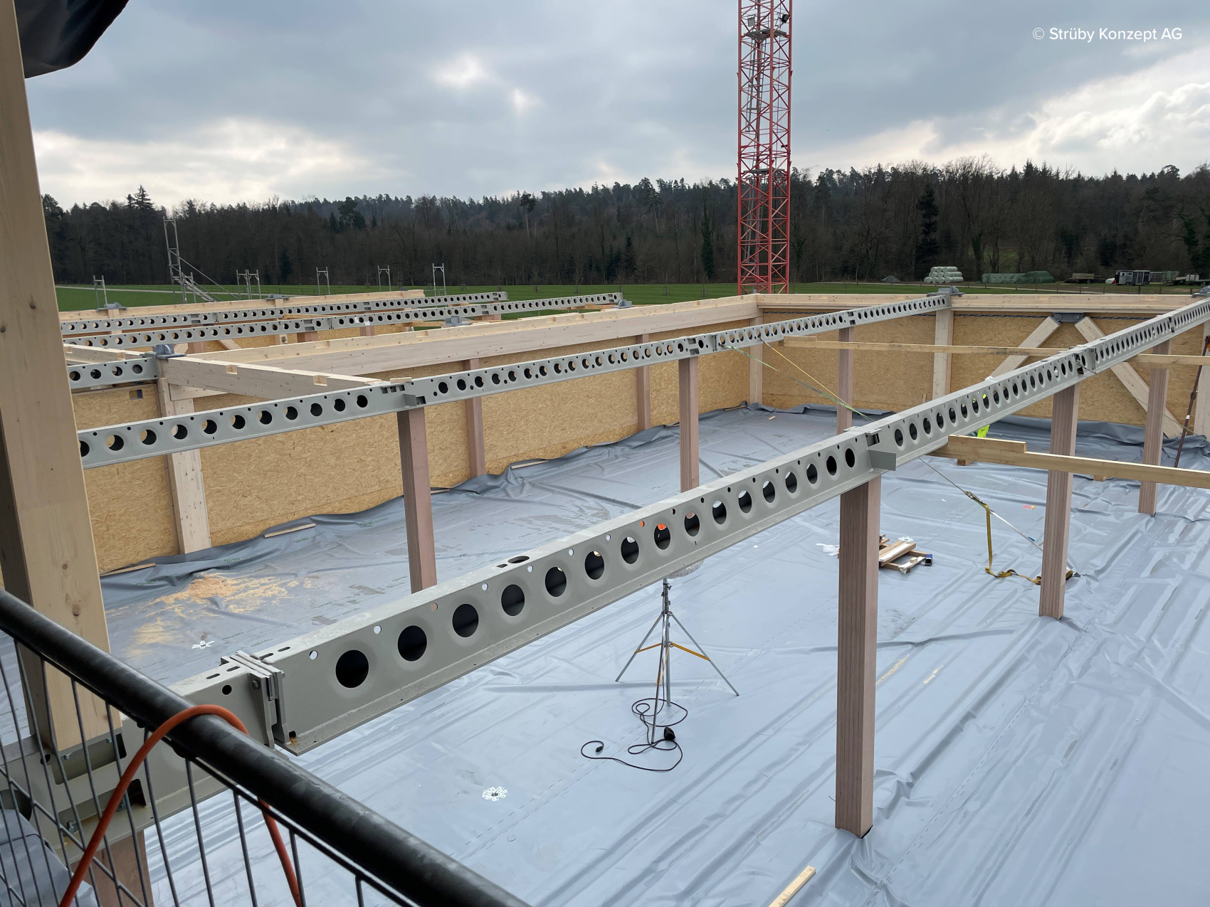 Un chantier de construction avec des poutres circulaires en acier, des murs en bois et un sol recouvert de plastique. En arrière-plan, on aperçoit une tour de grue rouge et des arbres sous un ciel nuageux.