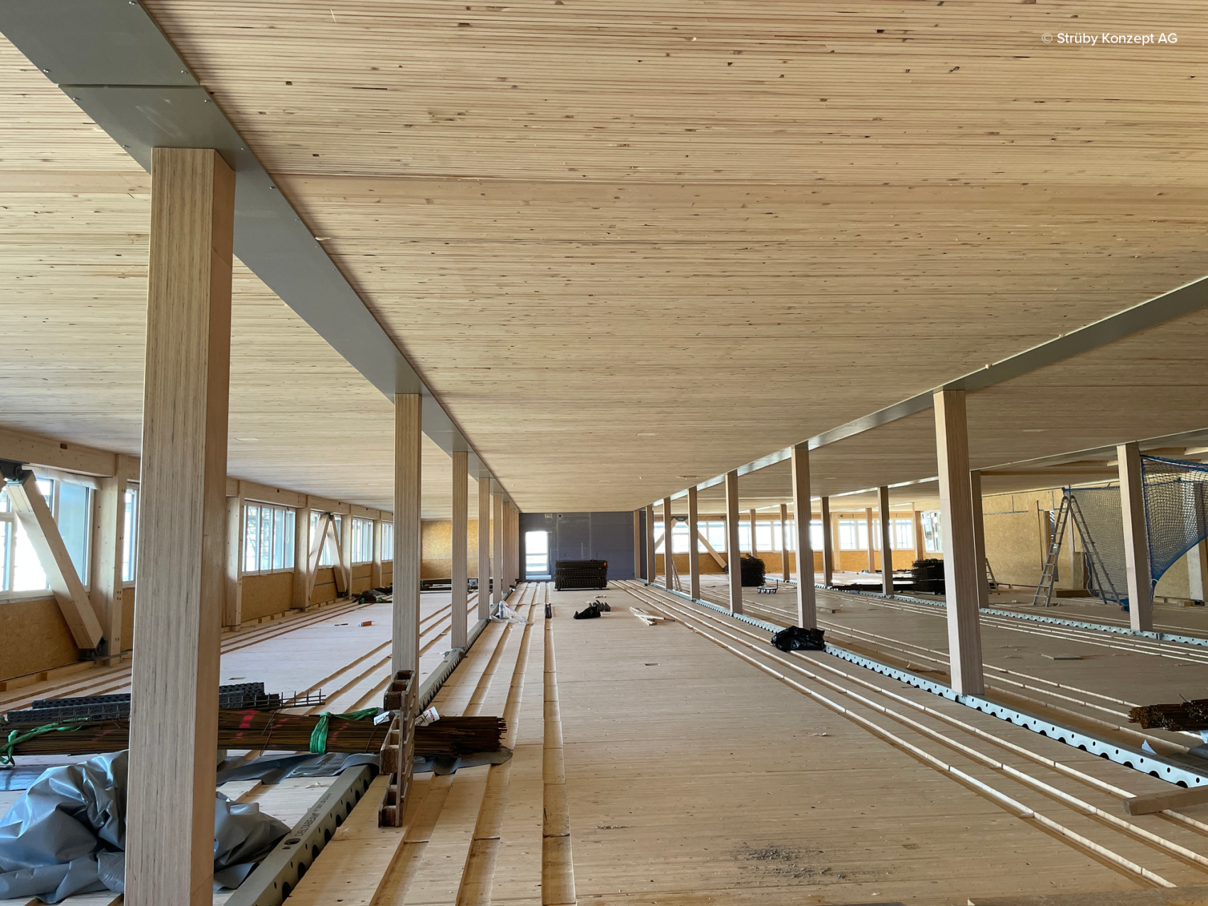 Un vaste intérieur ouvert en construction, avec parquet, plafonds et colonnes en bois. La lumière du jour entre par des rangées de fenêtres sur la gauche. Des matériaux et équipements de construction sont éparpillés au sol.